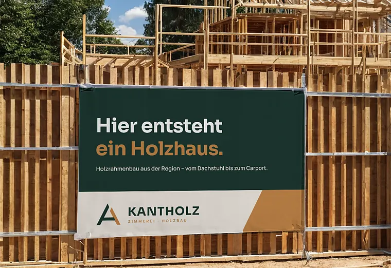 Bauzaunbanner „Hier entsteht ein Holzhaus“ vor einem Holzrohbau, Zimmerei Kantholz (Beispiel, fiktive Firma)