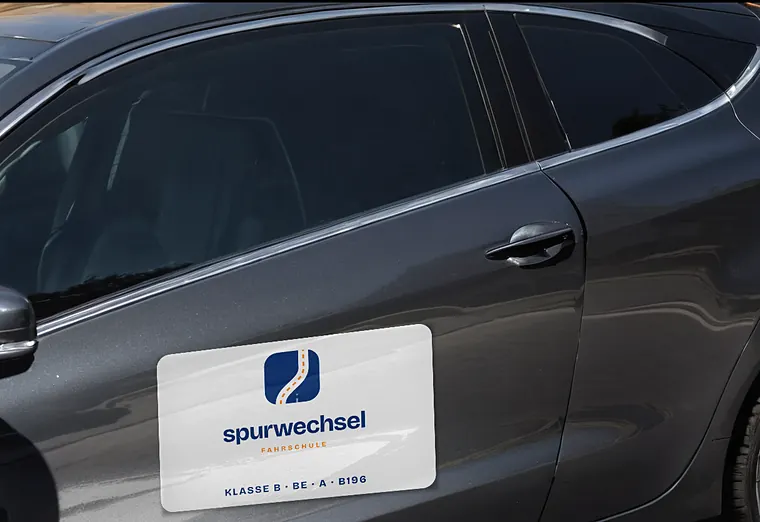 Magnetschild der Fahrschule Spurwechsel an einer dunklen Autotür (Beispiel, fiktive Firma)