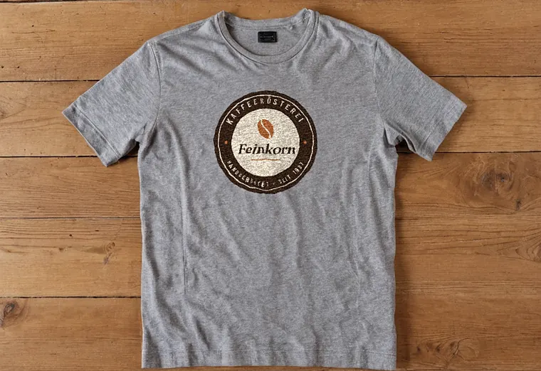 Graues T-Shirt mit Logo der Rösterei Feinkorn auf Holztisch (Beispiel, fiktive Firma)